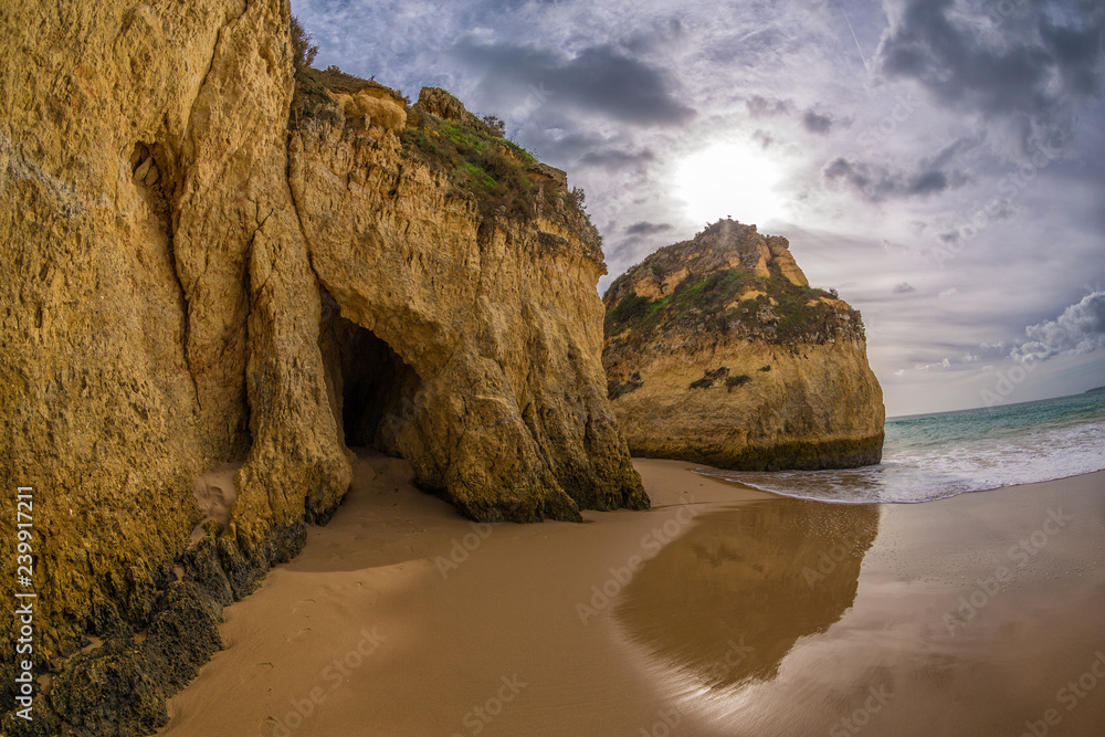 Famous rock formation with the cave on the beach of Tres Irmaos in Alvor, Portimão, Algarve, Portugal, Europe. Praia dos Tres Irmaos. The sun shines through the clouds under the rock, the waves