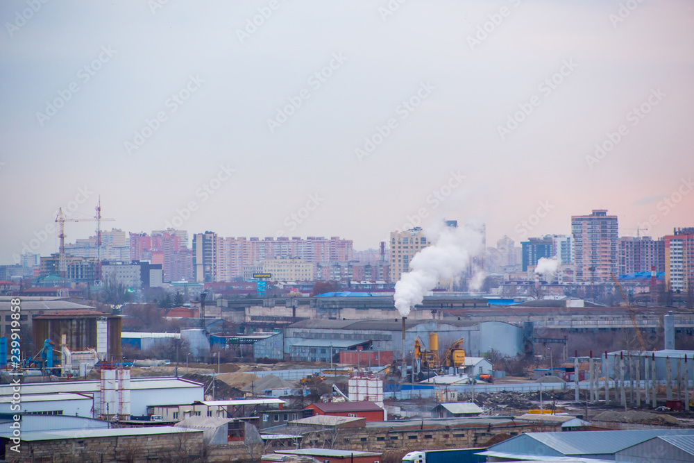 Landscape of evening city with high-rise buildings under construction, factories and pipes with smoke at sunset under boundless cloudy sky