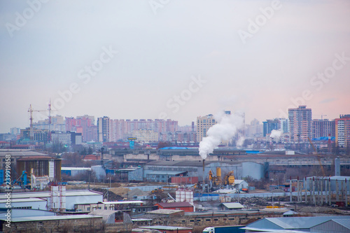 Landscape of evening city with high-rise buildings under construction  factories and pipes with smoke at sunset under boundless cloudy sky