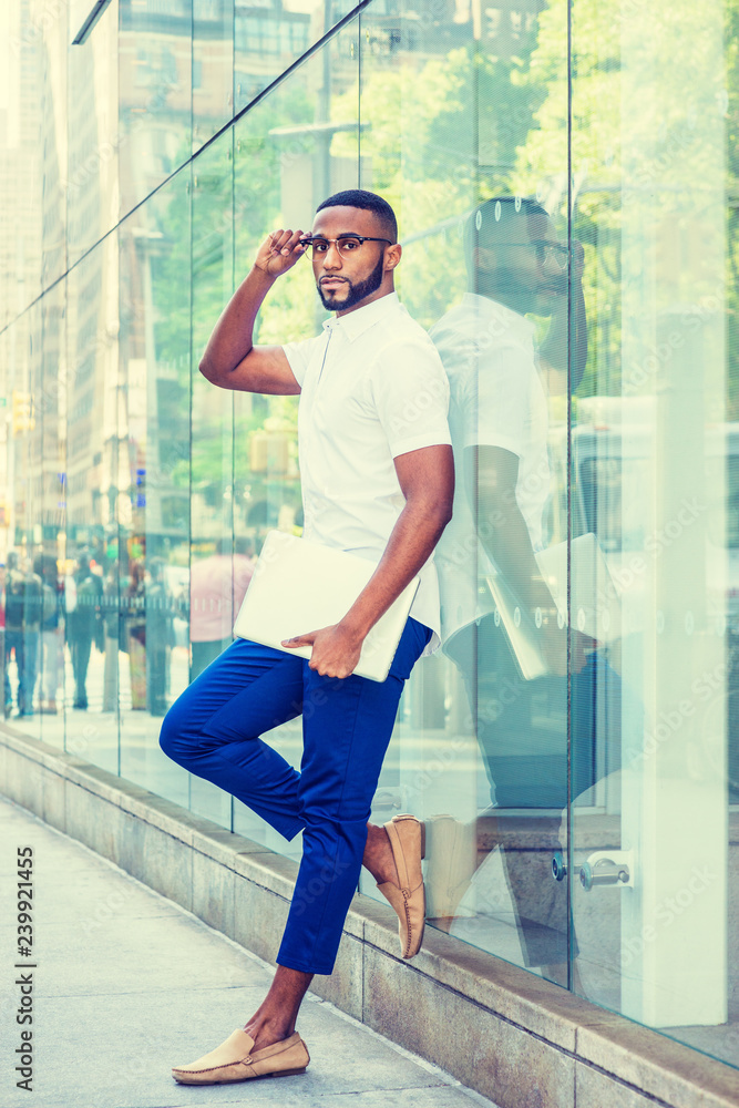 Young African American Man with beard, wearing white short sleeve shirt, blue pants, casual shoes, glasses, holding laptop computer, standing against glass wall on street in Manhattan, New York City..