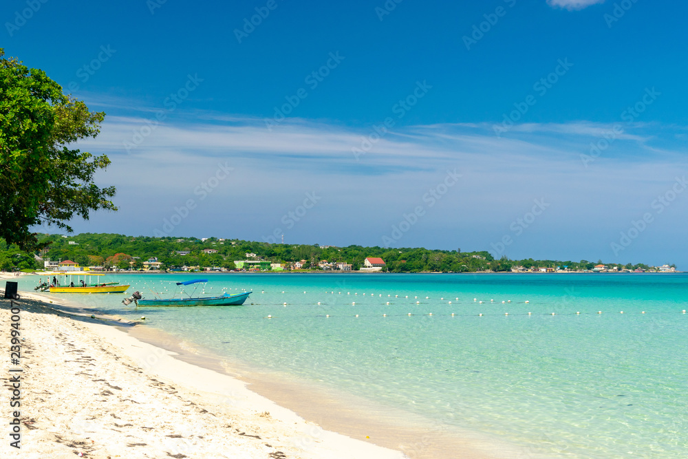Sunny day along the Seven Mile Beach in tropical Negril, Jamaica. Tour boats await passengers and caucasian tourists in the water at a distance.