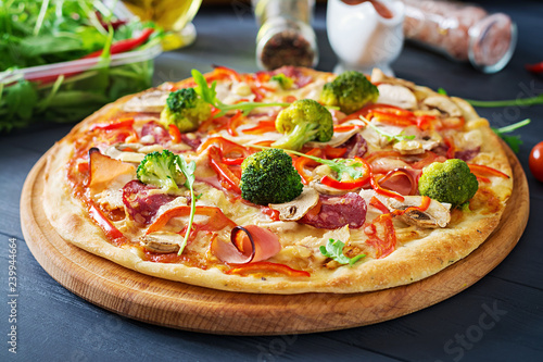Fresh italian pizza with chicken fillet, mushrooms, ham, salami, tomatoes, broccoli, cheese on on black background. Italian food.