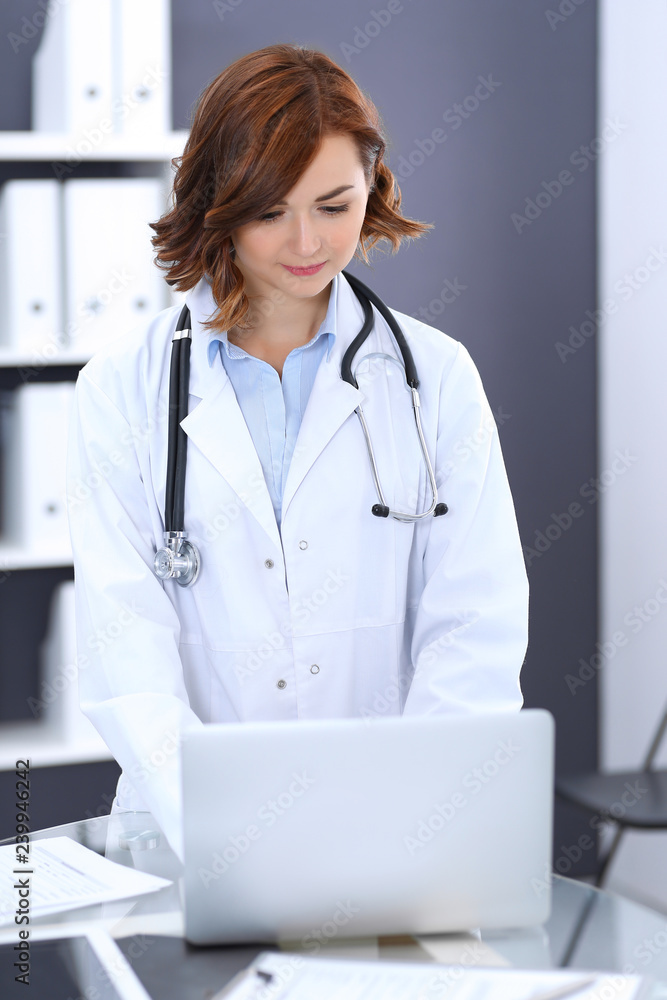 Happy doctor woman at work. Portrait of female physician using laptop computer while standing near reception desk at clinic or emergency hospital. Medicine concept Happy doctor woman at work. Portrait of female physician using laptop computer while standing near reception desk at clinic or emergency hospital. Medicine concept