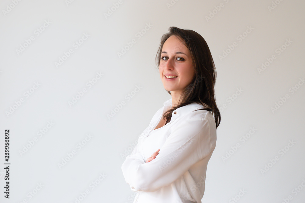 Smiling confident girl in white shirt posing with arms crossed. Side view of beautiful young woman turning face to camera. Side portrait concept Smiling confident girl in white shirt posing with arms crossed. Side view of beautiful young woman turning face to camera. Side portrait concept