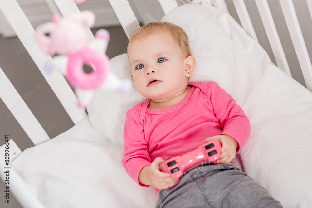 high angle view of adorable kid in pink shirt holding pink joystick and lying in crib high angle view of adorable kid in pink shirt holding pink joystick and lying in crib