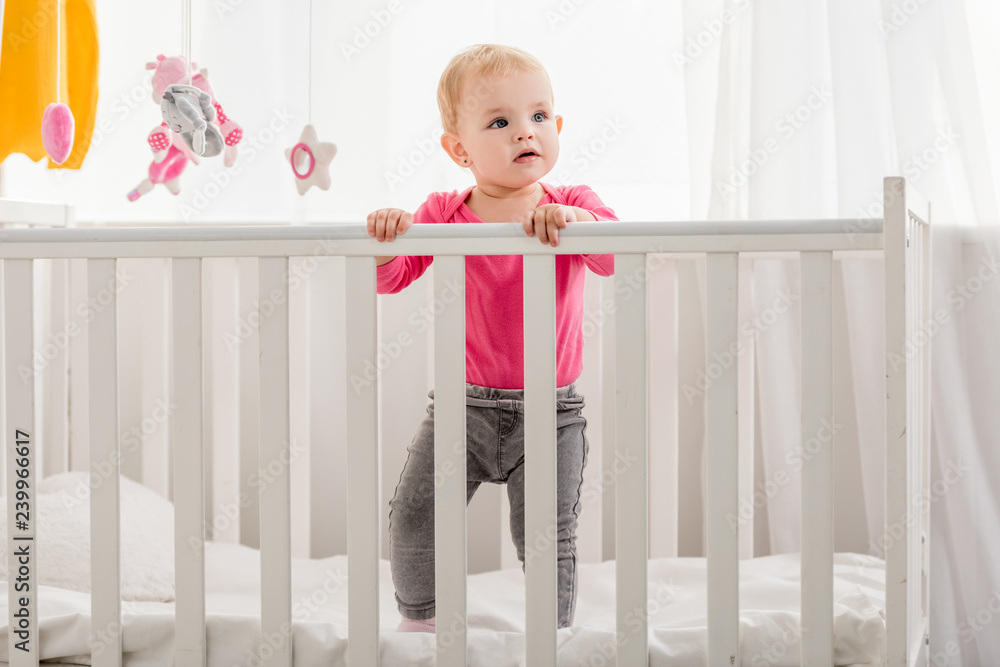 adorable kid in pink shirt standing in crib and looking away adorable kid in pink shirt standing in crib and looking away