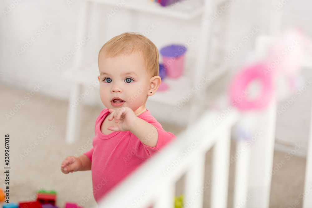 selective focus of adorable kid in pink shirt standing near crib in children room selective focus of adorable kid in pink shirt standing near crib in children room