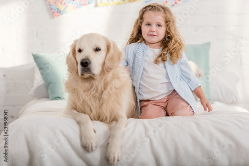 adorable kid and golden retriever dog sitting on bed in children room