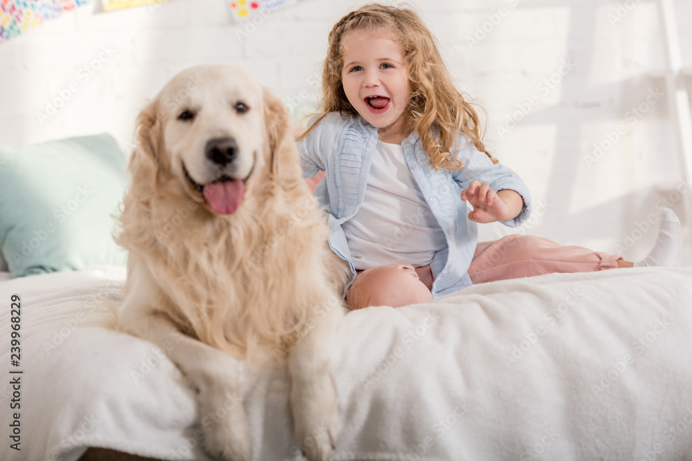 excited adorable kid and golden retriever sticking tongues out together on bed in children room