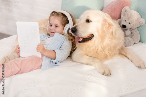 adorable child listening music with tablet and leaning on cute golden retriever on bed in children room