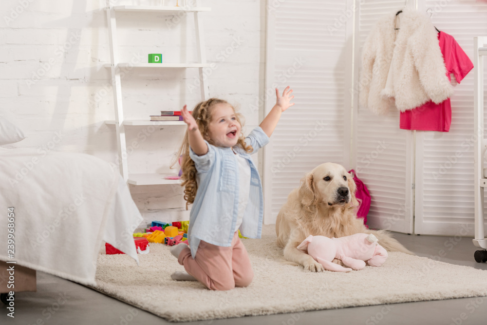 happy adorable kid standing on knees with open arms, golden retriever lying on carpet in children room happy adorable kid standing on knees with open arms, golden retriever lying on carpet in children room