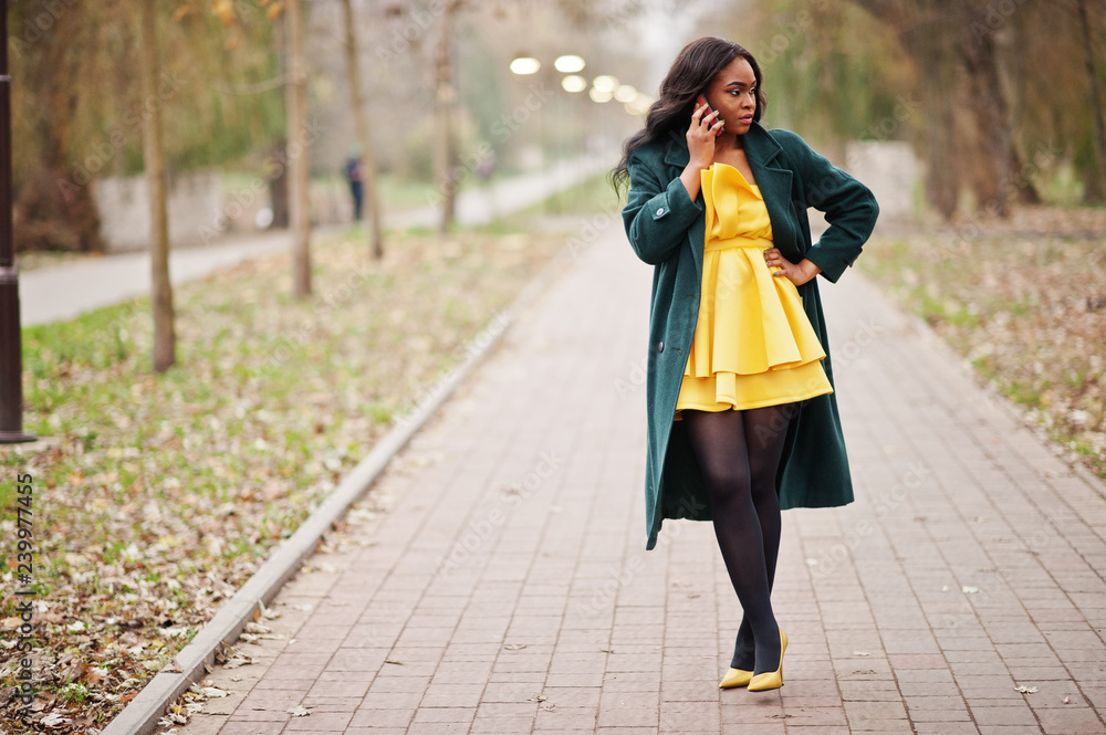 Stylish african american woman at green coat and yellow dress posed against autumn park and speaking on phone. Stylish african american woman at green coat and yellow dress posed against autumn park and speaking on phone.