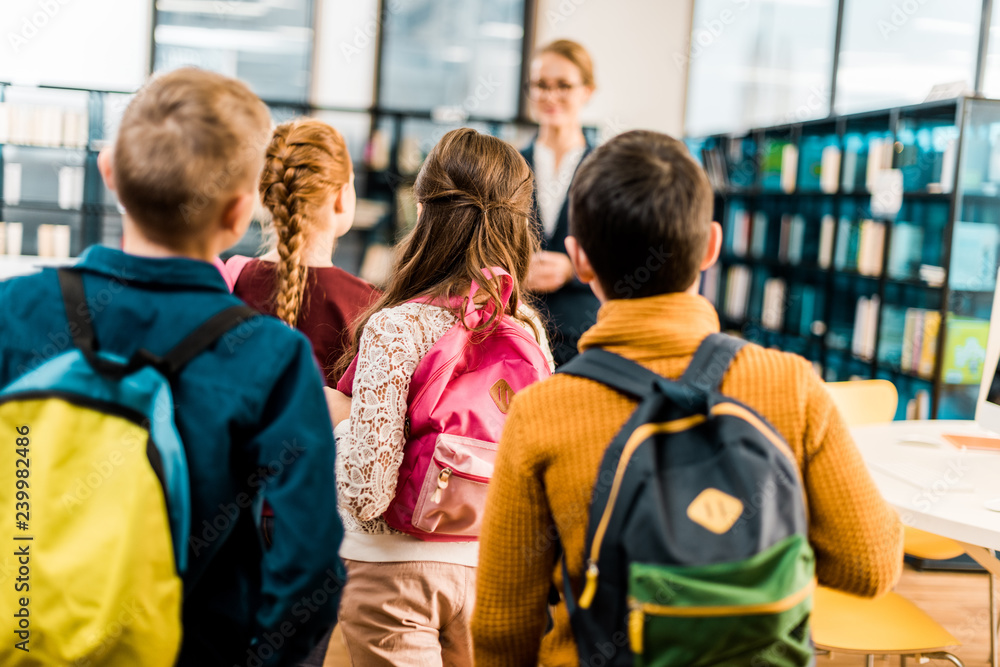 back view of schoolchildren with backpacks looking at librarian in library back view of schoolchildren with backpacks looking at librarian in library