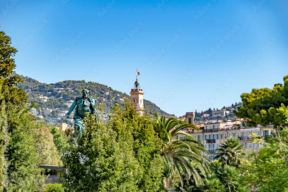 View over trees to historic buildings in Nice, France. In the foreground you can see a statue half hidden by trees.