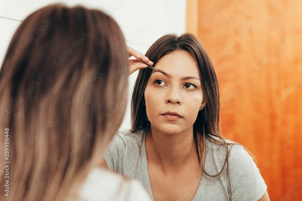 Young woman plucking eyebrow with tweezers in front of mirror Young woman plucking eyebrow with tweezers in front of mirror
