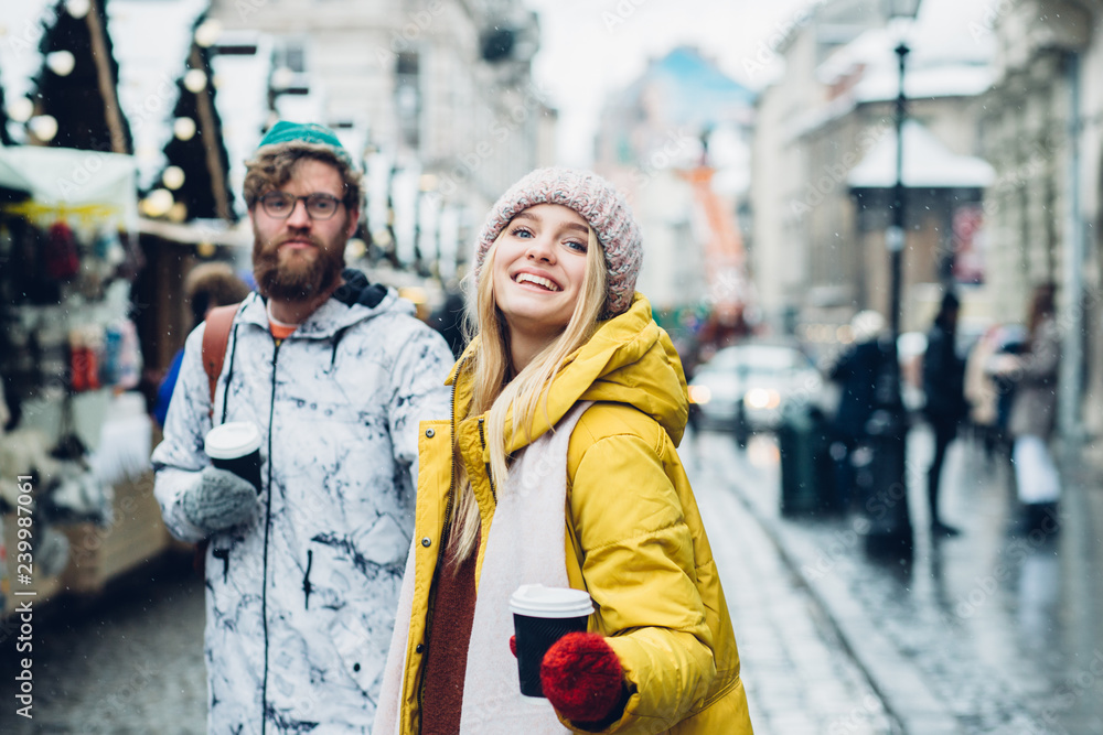 Young hupster couple in bright outwear doing christmas shopping at christmas fair, smiling, walking, drinking hot drinks in winter european city square, outdoor. Toned