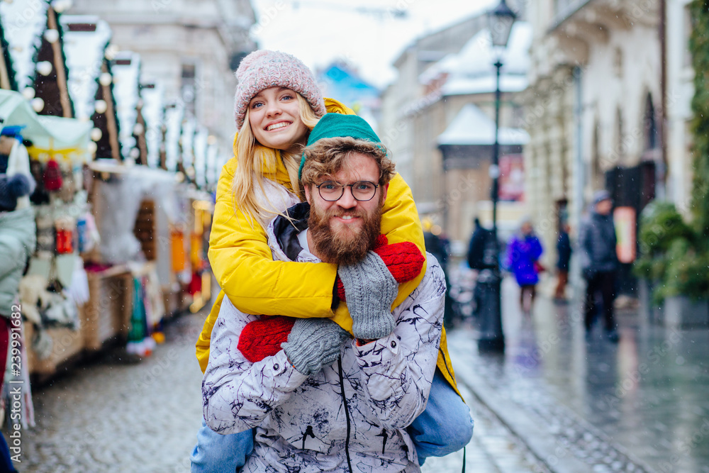 Couple in warm bright outwear, man giving piggyback to woman in street, outdoor. Handsome beard hipster man in eyeglasses and his girlfriend having fun while sightseeing in a foreign winter city.
