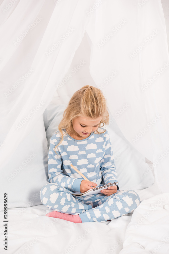 cute kid in pajamas sitting on bed with crossed legs and writing in notebook cute kid in pajamas sitting on bed with crossed legs and writing in notebook