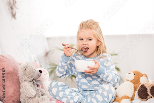 smiling child sitting in bed with soft toys and eating cornflakes