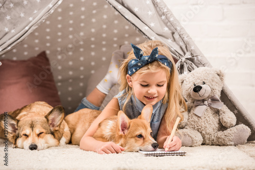 Wall Mural adorable child lying in wigwam with welsh corgi dogs and writing in notebook at