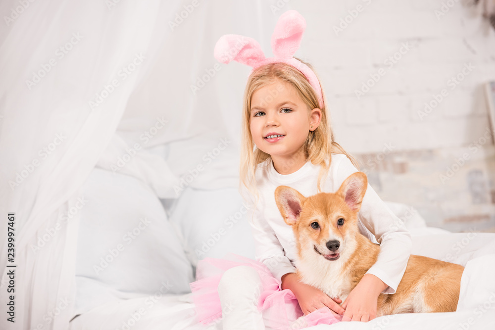 smiling child in bunny ears headband sitting with welsh corgi dog on bed at home