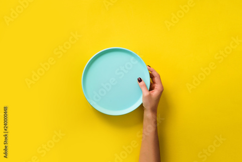 Female hand holding empty blue plate on yellow background with copy space. Healthy eating, dieting concept. Banner