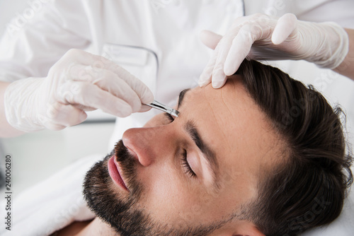 Calm bearded man undergoing plucking eyebrows procedure
