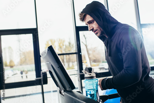 side view of handsome sportsman in hoodie exercising on treadmill in gym