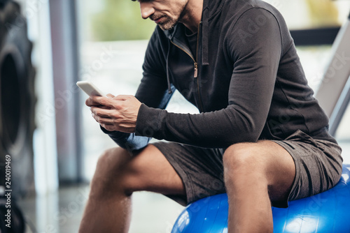 cropped image of sportsman sitting on fitness ball and using smartphone in gym