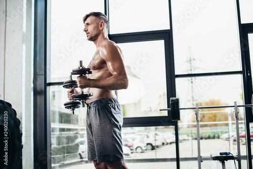 side view of handsome shirtless sportsman training with dumbbells in gym