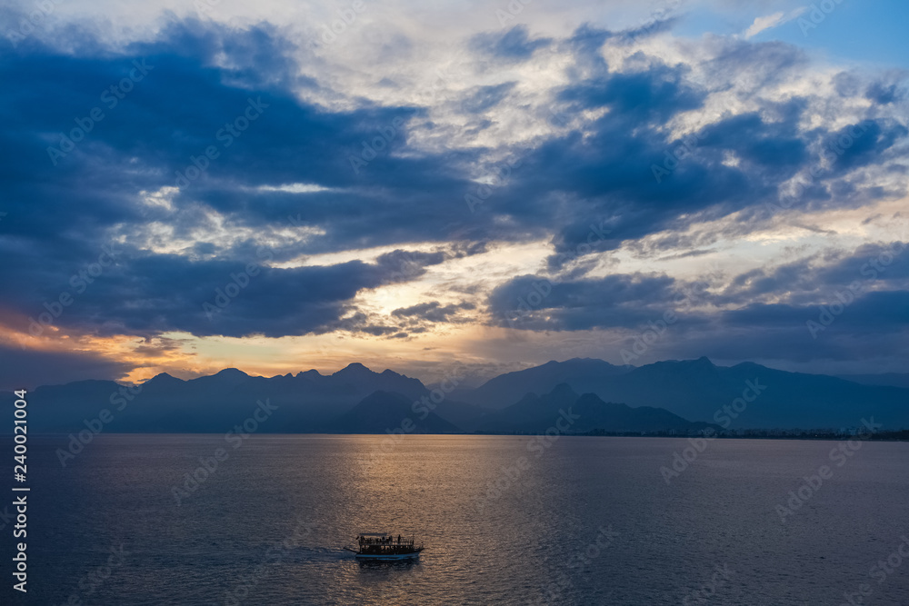 Tourists travelling by small ship in scenic marine background. Beautiful blue and gold sunset sunny sea water, mountains, cloudy fluffy sky, sailing yacht. Horizontal color photography.