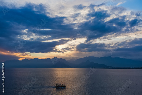 Tourists travelling by small ship in scenic marine background. Beautiful blue and gold sunset sunny sea water, mountains, cloudy fluffy sky, sailing yacht. Horizontal color photography.