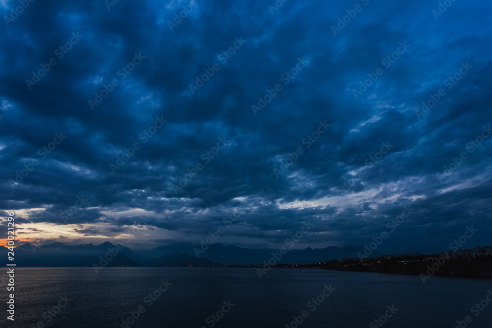 Beautiful sunset cloudy sky and dark sea water with silhouettes of mountains in background. Night is coming. Horizontal color photography.