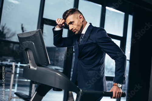 side view of tired handsome businessman in suit exercising on treadmill in gym photo