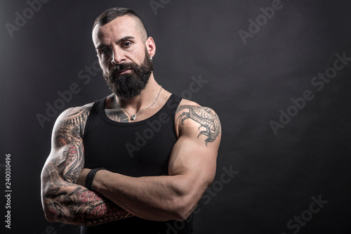 Uomo muscoloso e tatuato con braccia conserte guarda deciso in macchina - sfondo nero