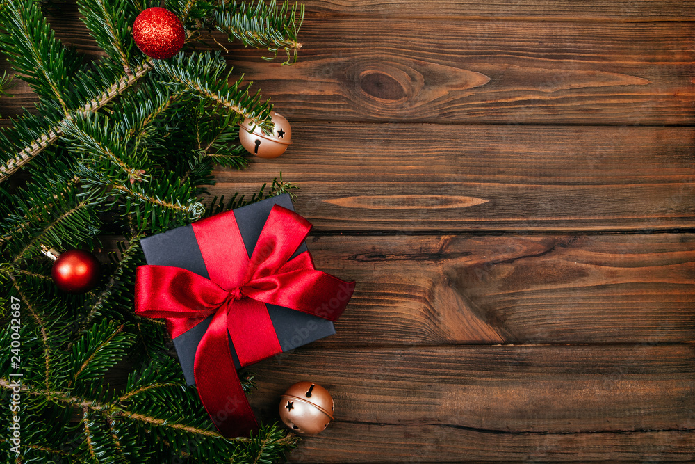Fototapeta premium Christmas gift box wrapped in kraft black paper with red ribbon decoration on rustic brown wooden background with fir tree, tree decoration and gold bells. Flatlay style.