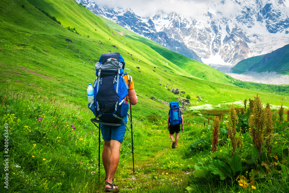Couple of young travelers hiking with backpacks on beautiful grassy trail in Alps mountains.
