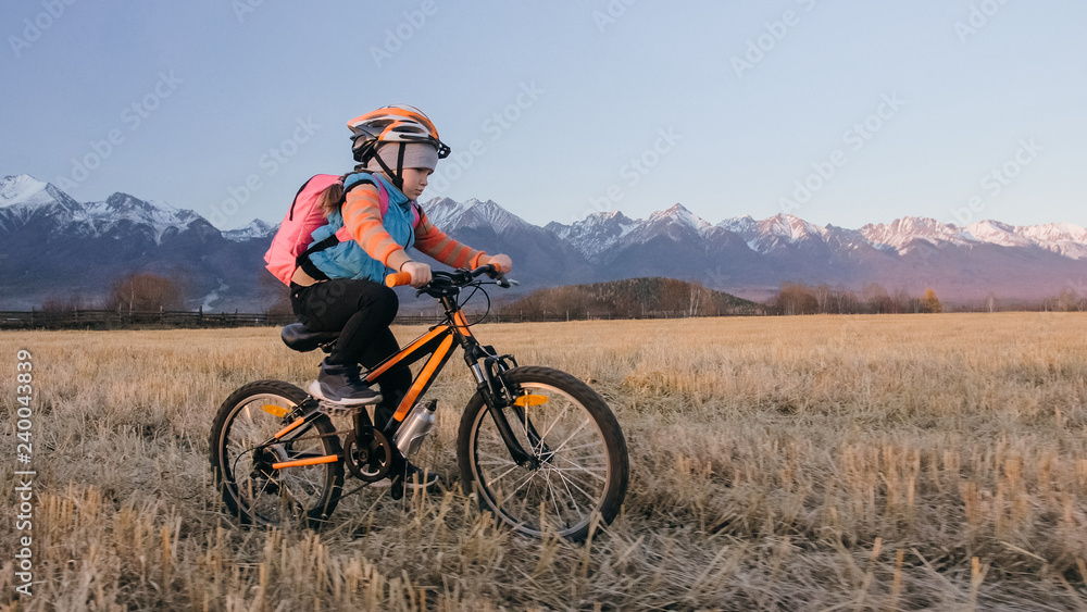 Obraz premium One caucasian children rides bike in wheat field. Little girl riding black orange cycle on background of beautiful snowy mountains. Biker motion ride with backpack, helmet. Mountain bike hardtail.