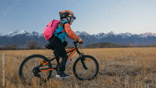 One caucasian children rides bike in wheat field. Little girl riding black orange cycle on background of beautiful snowy mountains. Biker motion ride with backpack, helmet. Mountain bike hardtail.