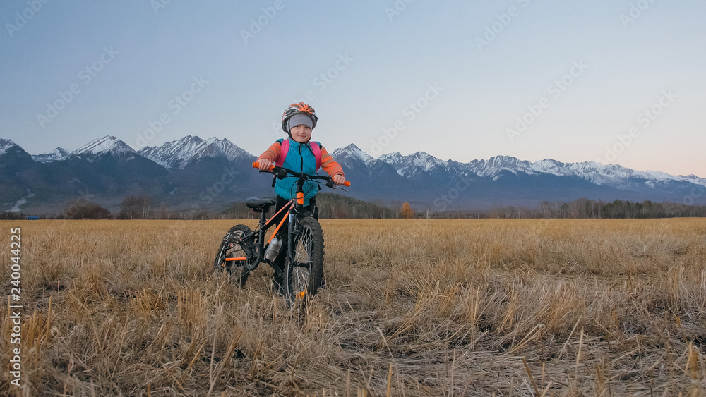 Obraz premium One caucasian children walk with bike in wheat field. Little girl walking black orange cycle on background of beautiful snowy mountains. Biker stand with backpack and helmet.