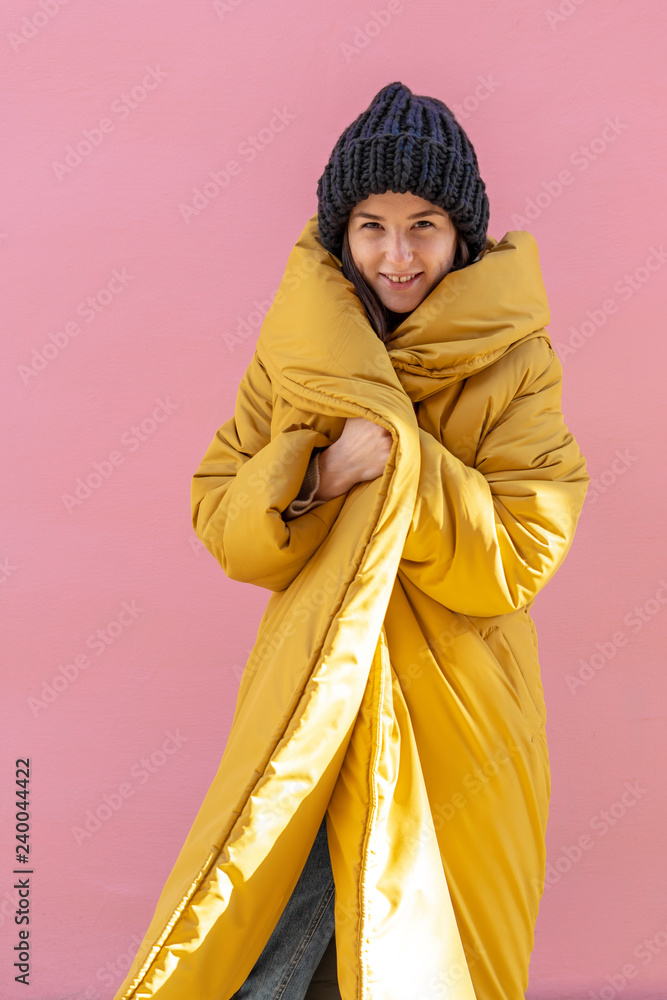 a stylish woman dressed in a knitted hat and a very warm and bright women's  winter jacket, a yellow jacket against a pink wall background Photos |  Adobe Stock