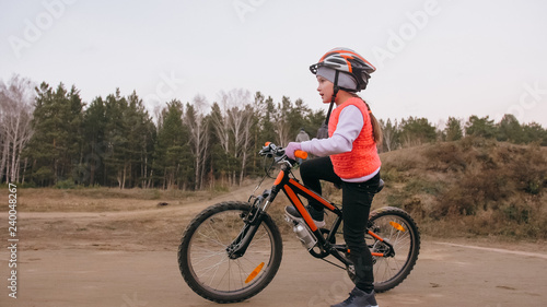 One caucasian children rides bike road track in dirt park. Girl riding black orange cycle in racetrack. Kid goes do bicycle sports. Biker motion ride with backpack and helmet. Mountain bike hardtail.