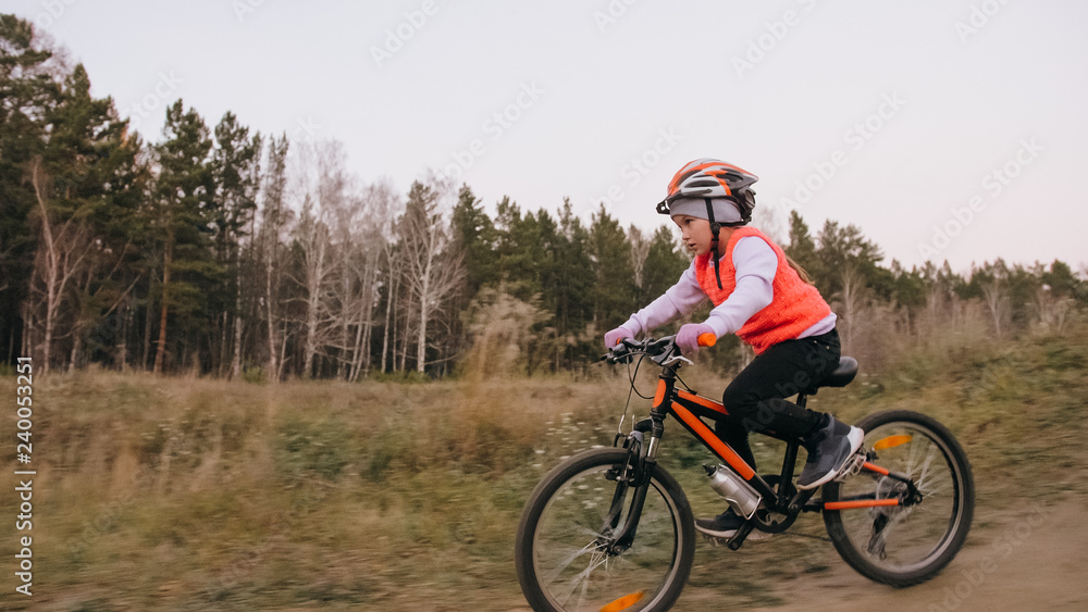 One caucasian children rides bike road track in dirt park. Girl riding black orange cycle in racetrack. Kid goes do bicycle sports. Biker motion ride with backpack and helmet. Mountain bike hardtail.