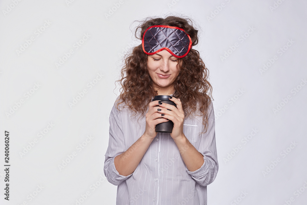 Φωτογραφία Shy pretty young woman with pleasant charming smile holds disposable cup with fresh coffee, dressed in casual stylish pajamas and sleeping mask, surprised and happy to start new day like this way