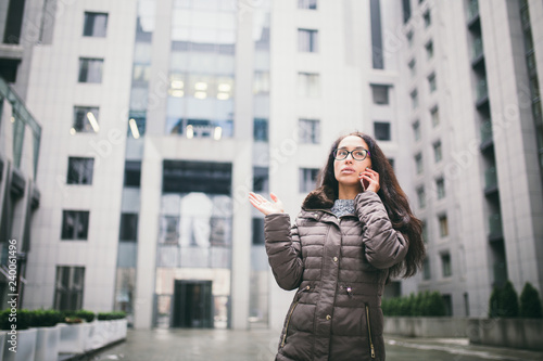 Theme is the business situation. Beautiful young woman of European ethnicity with long brunette hair wearing glasses and coat stands on background of business center and uses phone in hand near ear
