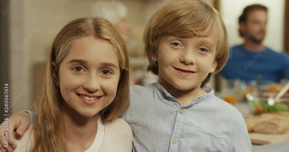 Portrait of the small caucasian sister and brother standing in front of the camera and smiling with a family at the dinner table behind. Close up. Indoor. Portrait of the small caucasian sister and brother standing in front of the camera and smiling with a family at the dinner table behind. Close up. Indoor.