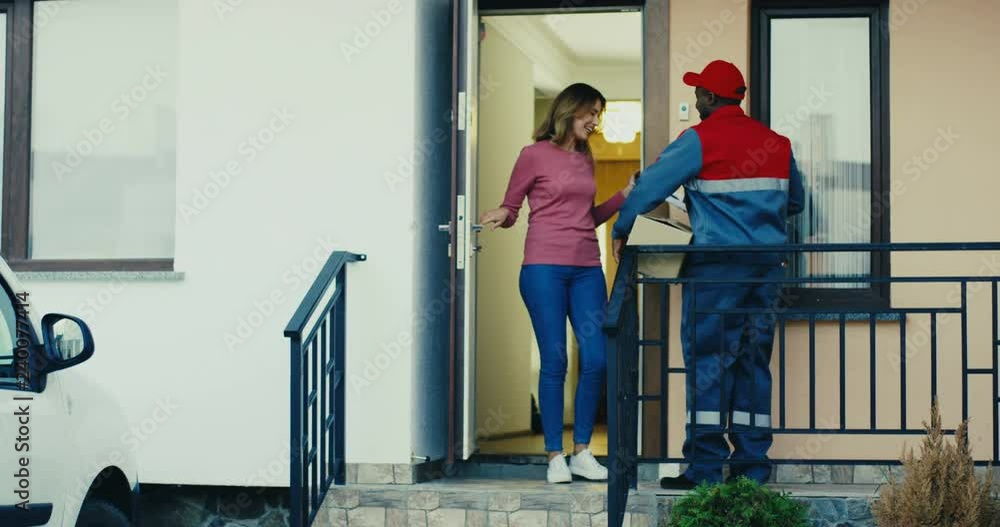 African American mailman in the uniform ringing in the doorbell, Caucasian woman opening a door and getting a parcel. Outside. African American mailman in the uniform ringing in the doorbell, Caucasian woman opening a door and getting a parcel. Outside.