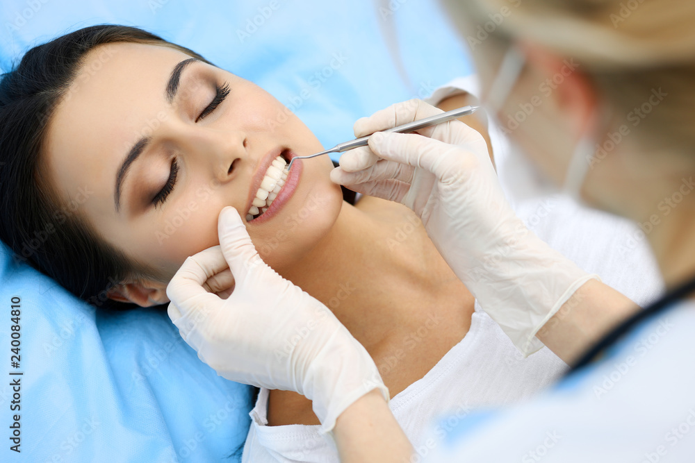 Naklejka premium Young female patient visiting dentist office.Beautiful woman with healthy straight white teeth sitting at dental chair with open mouth during oral checkup while doctor working at teeth