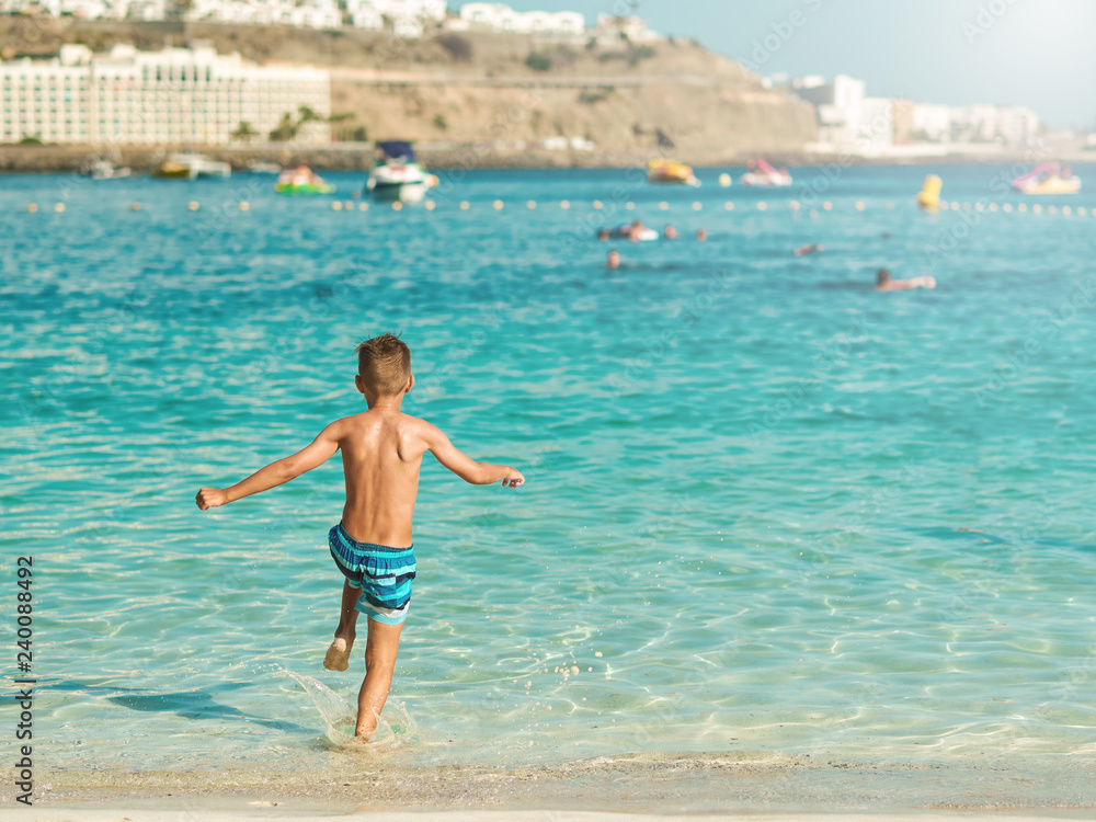 Caucasian boy in a striped swimming shorts is running towards the ocean. He is holding his hands wide open.