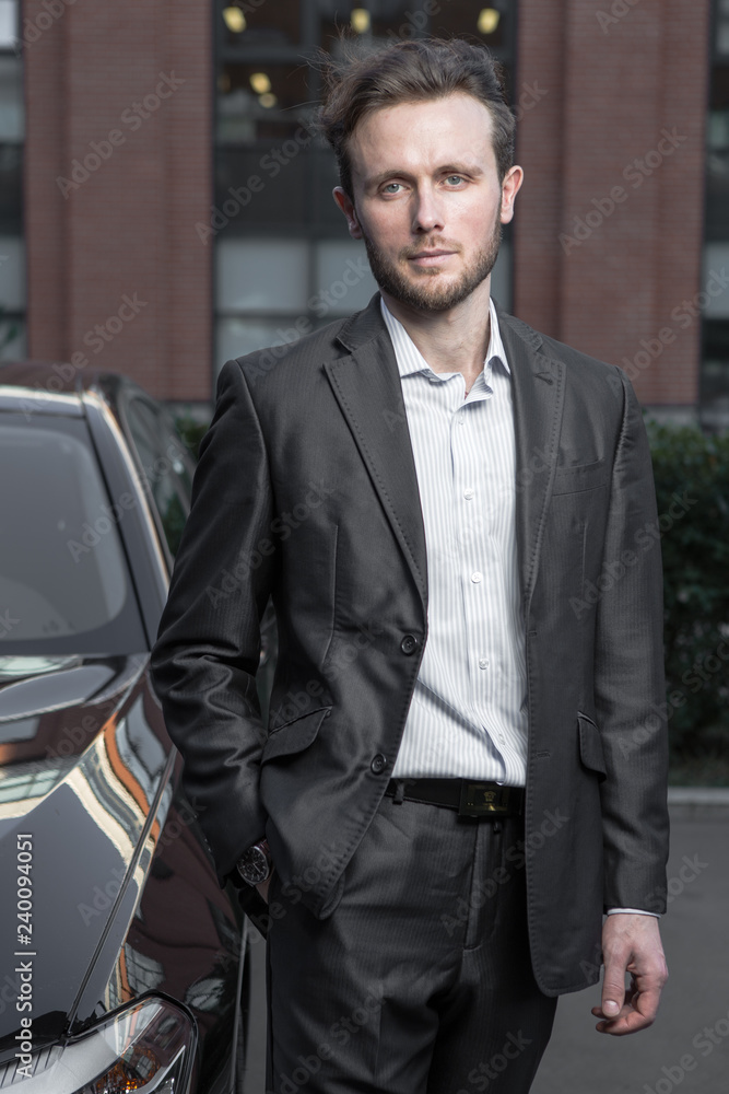 Attractive successful young businessman in a business suit near his car premium class car and office buildings in background